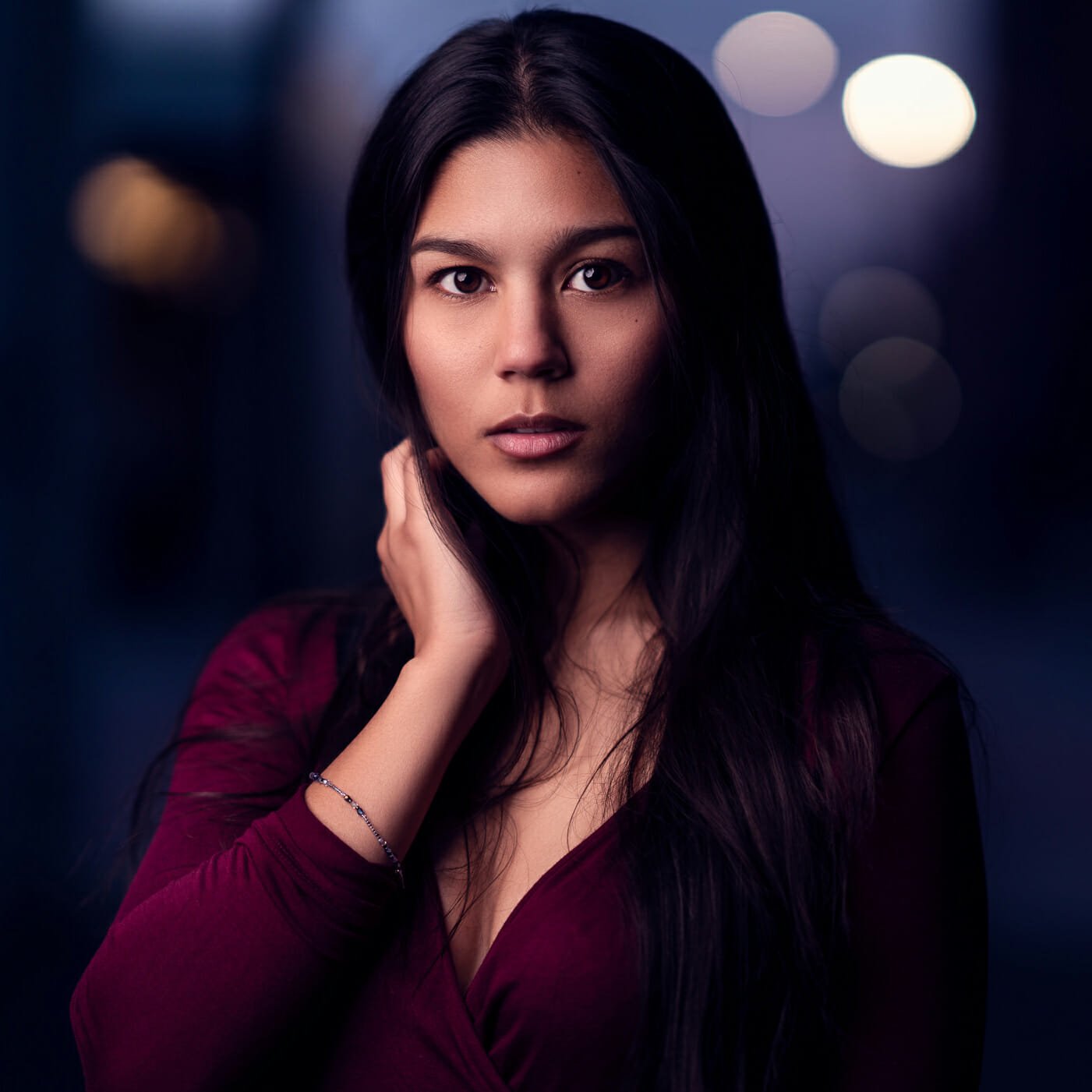Morgan at night, with a red top. She is bringing up her right hand to gently touch the back of her neck. One light setup with the light to camera left for Rembrandt lighting.