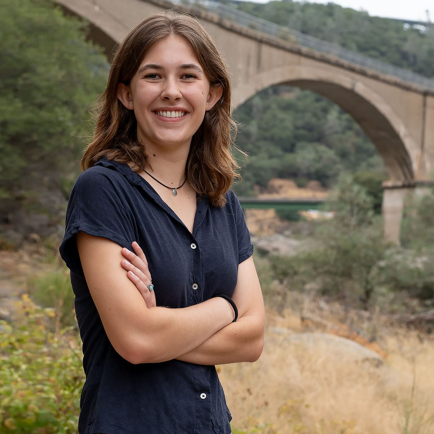 A senior picture of a girl wearing a blue shirt and crossing her arms. The picture is taken in front of No Hands Bridge in Auburn CA.